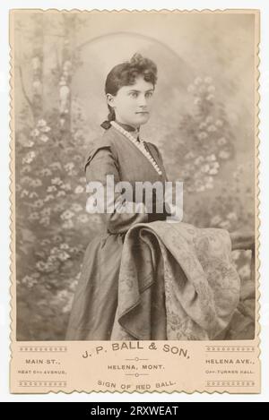 Eine Schwarzweißfotografie einer Frau, die im J. P. Ball & Son Studio in Helena, MT, fotografiert wurde. Das Foto ist auf einer hellen Schrankkarte mit dekorativen Kanten montiert und der Name und die Position des Fotostudios ist auf der Vorderseite entlang der Unterseite aufgedruckt. Die Frau steht im Profil von 3/4 und posiert vor einer floralen Waldkulisse. Sie hat dunkle Haare, die zurückgezogen wurden, und trägt ein dunkelfarbenes Kleid mit weißem Kragen. Ihr rechter Arm ruht auf einem mit Textil bedeckten Zaun. Stockfoto