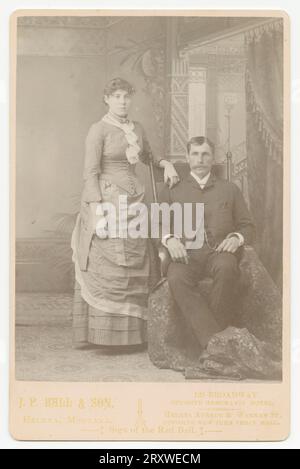 Ein Schwarzweißfoto von einem Mann und einer Frau, das im J. P. Ball & Son Studio in Helena, MT, fotografiert wurde. Das Foto ist auf einer hellen Schrankkarte montiert, auf der der Name und die Position des Fotostudios auf der Vorderseite entlang der Unterseite aufgedruckt sind. Die Frau trägt ein helles Kleid mit einem gerafften Halsteil und fingerlosen weißen Handschuhen. Ihre linke Hand wird über die Rückseite eines Stuhls gelegt, in dem der Mann sitzt. Er hat einen Schnurrbart und trägt einen dunklen Anzug. Unter ihm ist ein langes Tuch, das vor seinen Füßen zusammengebunden ist. Sie sind vor einem bemalten inneren Hintergrund aufgestellt. Stockfoto