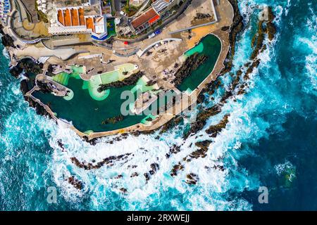 Aus der Vogelperspektive sehen die Menschen, die in den natürlichen Lavapools in Porto Moniz, Madeira, Portugal schwimmen, während das Meer um sie herum tobt und die Gehwege überflutet Stockfoto