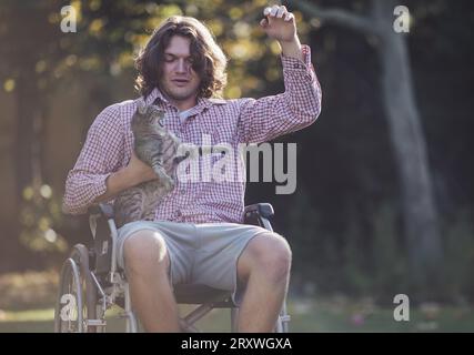 Junger positiver Mann im Rollstuhl, der mit Katze im Park spielt Stockfoto