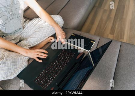 Eine junge Frau im Schlafanzug sitzt auf der Couch und benutzt einen Laptop Stockfoto