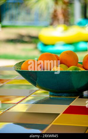 Eine Schüssel voller saftiger Orangenfrüchte auf einem vielfarbigen Tisch. Orangen sind bereit, serviert zu werden. Wunderschöner Garten im Hintergrund an einem sonnigen Tag. Stockfoto