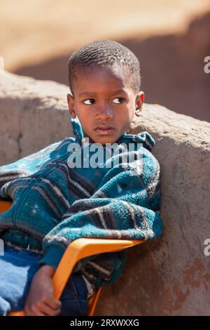 afrikanisches Kind im Dorf, das eine Jacke trug, saß auf dem Stuhl im Hof in der Schule Stockfoto