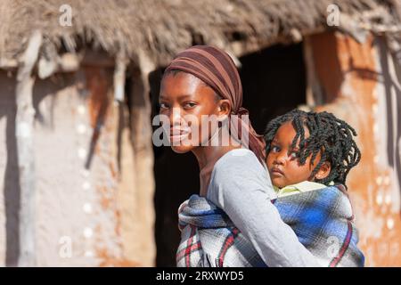 die afrikanische Mutter im Dorf trägt ihr Kind auf traditionelle Weise in einer Decke im Hintergrund mit der Schlammhütte im Rücken Stockfoto