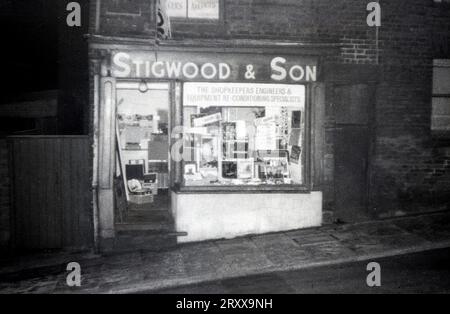 1960er Jahre, historisch, an einer schrägen Straße, das Äußere des Stigwood & Son Stores, Fensterbanner mit der Aufschrift „The Shopkeepers Engineers & Equipment Re-Conditioning Specialists, Oldham, England, Großbritannien. Die Fensteranzeige enthält Schallplatten, Plattendecks, Funkgeräte + Poster Abour Waagen, Gewichtungsmaschinen und Slicer. Stockfoto