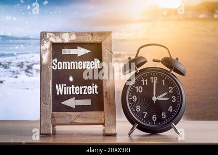 26. September 2023: Tafel mit der Aufschrift Sommerzeit und Winterzeit vor dem Himmel mit Sonnenschein und Schnee mit einem schwarzen Wecker. Zeitwechsel-Symbol Bild FOTO-MONTAGE *** Tafel mit der Aufschrift Sommerzeit und Winterzeit vor Himmel mit Sonnenschein und Schnee mit einem schwarzen Wecker. Zeitumstellung Symbolbild FOTOMONTAGE Stockfoto