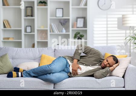 Ein junger muslimischer Mann liegt zu Hause auf der Couch, krank und hält seinen Bauch mit der Hand, leidet unter Schmerzen, Übelkeit, Unwohlsein. Stockfoto