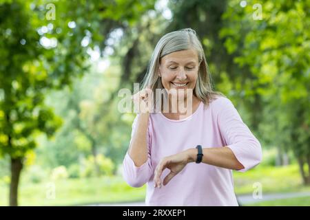 Ältere grauhaarige schöne Frau läuft und macht Sport im Park und überprüft die Zeit auf dem Tracker, glücklich mit dem Ergebnis. Er schaut auf die Smartwatch am Handgelenk. Stockfoto