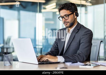 Ein junger indischer Geschäftsmann arbeitet im Büro auf einem Laptop und trägt Kopfhörer. Konzentriertes Tippen auf der Tastatur, Chats. Stockfoto