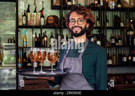 Vorderansicht eines fröhlichen Kellners, der auf die Kamera schaut und lächelt, während er ein Tablett mit Bieren hält. Stockfoto
