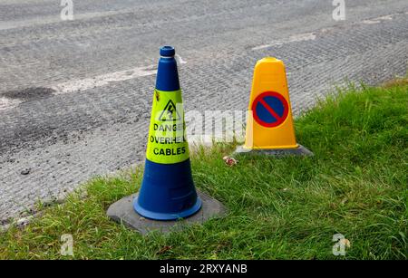 Blaue und gelbe Verkehrskegel mit Warnschildern auf dem Gras am Straßenrand in der Nähe einer asphaltierten Straße, die auf Gefahren und Parkbeschränkungen hinweisen Stockfoto