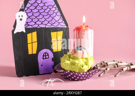 Komposition mit köstlichem Halloween-Cupcake, Papierhaus, brennender Kerze und Skeletthand auf rosa Hintergrund, Nahaufnahme Stockfoto