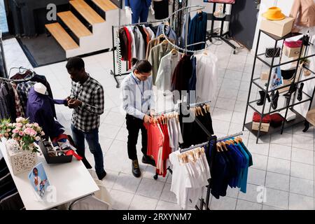 Asiatischer Mitarbeiter bereitet Boutique für die Eröffnung vor und arrangiert trendige Waren auf Kleiderbügeln im Einkaufszentrum. Mann trägt formelles Hemd und sieht sich die Regale mit der neuen Modesammlung im Bekleidungsgeschäft an Stockfoto