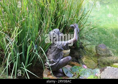 Eine Bronzeskulptur eines kleinen Jungen, der einen Frosch am Ende eines Teichs zwischen Wasserpflanzen in den Na Aina Kai Botanical Gardens in Kauai, Hawaii, USA hält Stockfoto