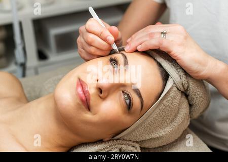 Große inhaltliche Dame mit Handtuch auf dem Kopf, die auf dem Bett liegt, während anonyme Kosmetikerin im Schönheitssalon Augenbrauen zupft Stockfoto