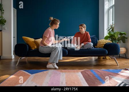 Aufgebrachtes Teenager-Mädchen, das auf dem Sofa sitzt und zuhört, wie die wütende Mutter sie wegen schlechter Schulnoten schimpft Stockfoto