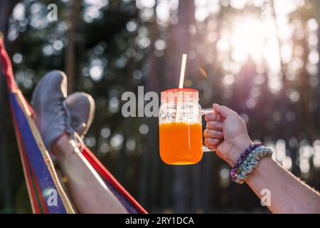 Orange Smoothie oder Saft im Trinkglas. Frau, die sich in der Hängematte im Wald entspannt, während der Sonnenuntergang Stockfoto