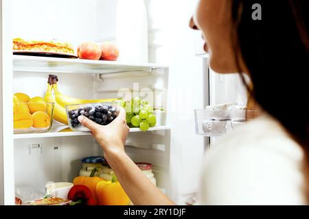Frau, die eine Schachtel Blaubeere aus dem offenen Kühlschrankregal oder der Kühlschublade mit Obst, Gemüse, Bananen, Pfirsichen nimmt, schnappt oder nimmt, Stockfoto