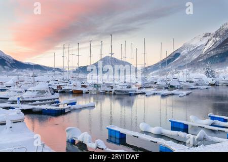 Ein ruhiger Wintermorgen offenbart einen Jachthafen, der mit Schnee bedeckt ist, mit Booten, die friedlich vor der Kulisse der Berge und des sanften Pastellhimmels verankert sind. Stockfoto