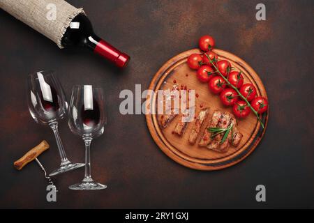 In Scheiben geschnittene gegrillte Schweinesteaks mit einer Flasche Wein, Weinglas, Korkenzieher, Kirschtomaten und Rosmarin auf rostigem Hintergrund. Draufsicht. Stockfoto