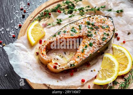 Gebackener Lachs mit aromatischen Kräutern und Zitrone auf Backpapier, Draufsicht. Gebratenes Lachssteak mit Thymian und Zitrone auf Holzteller auf schwarzem Tisch. Stockfoto