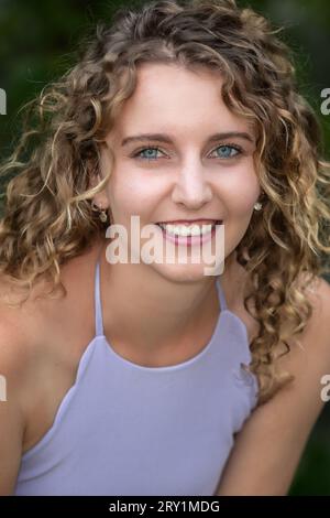 Kopf-Schulter-Porträt einer lächelnden, jungen Kaukasierin mit lockigem blondem Haar und blauen Augen, draußen im Sommer Stockfoto