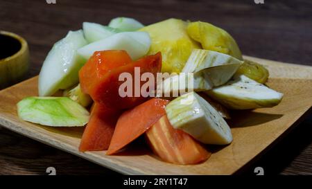 Rujak-Buah oder Obstsalat mit Pfirsichsoße, serviert auf einem Holzteller. Papaya, Mango, Banane, Ananas und Gurke. Stockfoto