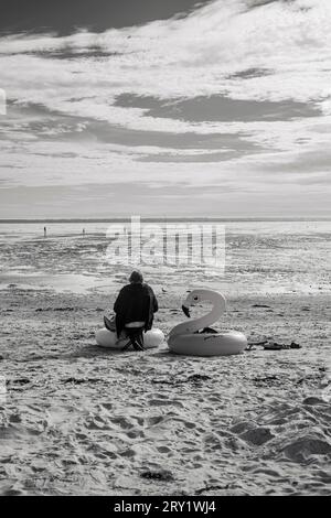 Frankreich, Les Moutiers-en-Retz, 44, ältere Frau, die allein neben einer Kinderboje in der Mitte eines leeren Strandes sitzt, Sommer 2022. Stockfoto