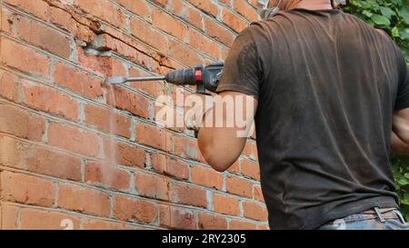 Ein Baumeister in einem schwarzen T-Shirt mit einem Bohrer in den Händen höhlt eine Schicht roten Ziegels an der Außenwand eines Hauses aus, um eine Fensteröffnung zu bilden Stockfoto