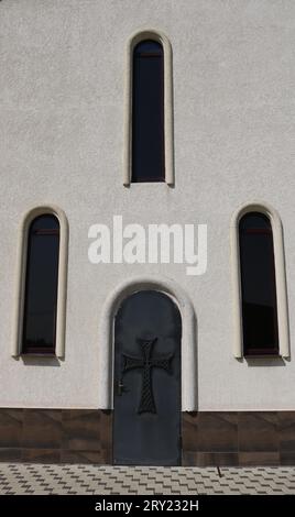 Helle Wand eines religiösen Gebäudes mit bogenförmigen, schmalen Fenstern und einer alten Tür mit einem christlichen Kreuz, vertikalem Bild, Fragment der Außenseite Stockfoto