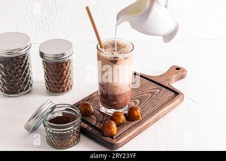 Ein Milchstrom aus einer Kanne wird in ein hohes Glas eiskalten Kaffees gegossen. Dosen zur Aufbewahrung von Kaffee. Zubereitung eines köstlichen Getränks. Stockfoto