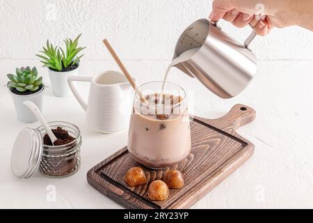 Ein Milchstrom aus einer Metallkanne wird in ein großes Glas eiskalten Kaffees gegossen. Dosen zur Aufbewahrung von Kaffee. Zubereitung eines köstlichen Getränks. Stockfoto
