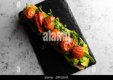 Draufsicht auf appetitliche, saftige, gereifte rote Tomaten, die auf Avocadoscheiben, Kräutern, grünen Blättern auf schwarzer Oberfläche platziert werden, während sie auf Marmorplatte in d serviert werden Stockfoto