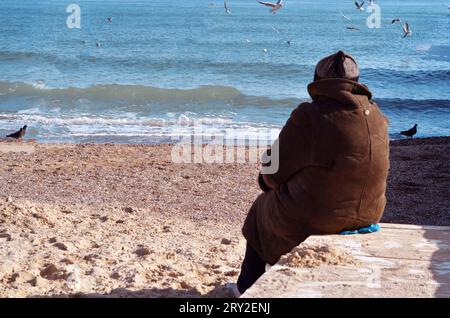 Eine Person, die auf einem Felsvorsprung sitzt und auf das Meer blickt. Dieses Bild zeigt eine ruhige Outdoor-Szene an einem Strand. Stockfoto