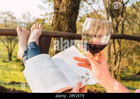 Gesichtslose Frau mit einem Glas Rotwein beim Lesen des Buches und beim Entspannen auf einer Veranda in der Natur Stockfoto