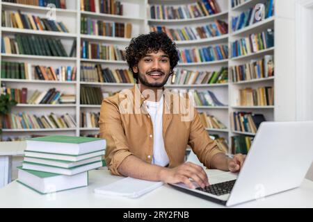 Porträt eines lächelnden jungen indischen Mannes, der in einer Bibliothek am Laptop studiert und arbeitet. An einem Tisch mit Büchern sitzen und auf die Kamera schauen. Stockfoto