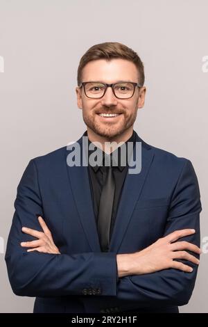 Glücklicher erfolgreicher, selbstbewusster Geschäftsmann im Anzug mit überkreuzten Armen auf der Brust, der lächelnd die Kamera anschaut Stockfoto