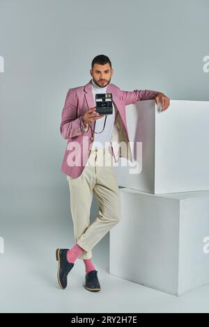 Stylischer Mann in einem lilafarbenen Blazer und einer weißen Hose, der Fotos auf einer Vintage-Kamera in der Nähe von Würfeln auf Grau macht Stockfoto