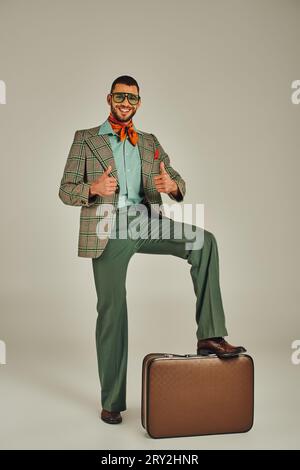 Glücklicher Mann in Retro-Kleidung und Sonnenbrille mit Daumen nach oben in der Nähe von Vintage-Koffer auf Grau Stockfoto