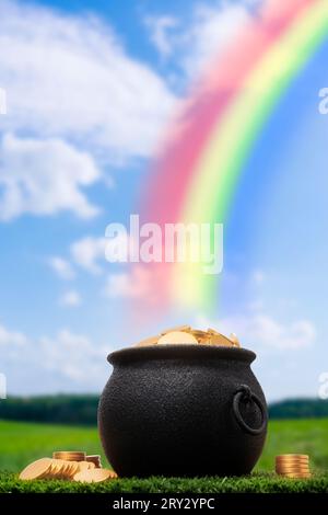 Ein schwarzer gusseiserner Topf voller Goldmünzen, der im Gras am Ende eines Regenbogens sitzt. Einige Goldmünzen sind auf dem Gras neben dem Topf gestapelt. Stockfoto