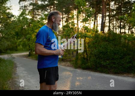 Pensionierter Jogger macht Pause, trinkt Wasser und schaut sich lustige Videos am Telefon an Stockfoto