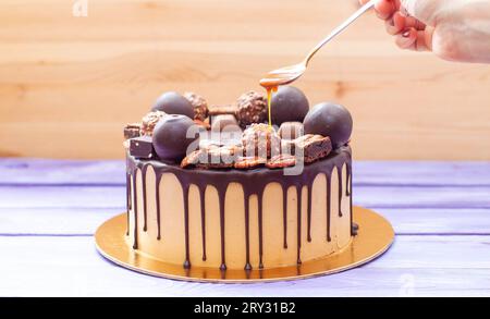Die Hand der Frau gießt gesalzene Karamellsauce auf den Schokoladenkuchen, dekoriert mit Süßigkeiten, Trüffeln, Keksen und Nüssen Stockfoto