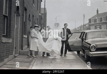 1960er Jahre, historisch, verlassen ein kleines Reihenhaus, mit ihrer Familie hilft, eine junge Braut in einem Hochzeitskleid und Schleier auf dem Bürgersteig, kurz vor dem Einsteigen in das Hochzeitsauto, ein amerikanischer Ford, geparkt auf der Ripponden Rd, Oldham, England, Großbritannien. Das Bulls Head Public House kann man die Straße hinauf sehen. Stockfoto