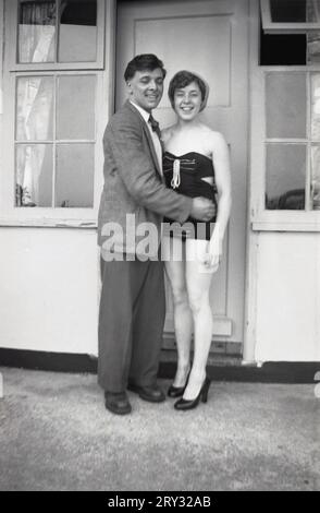 1960er Jahre, historisch, ein glückliches junges Paar, das vor ihrem Ferienhaus für ein Foto steht, die junge Frau im Badeanzug der Ära, England, Großbritannien. Stockfoto