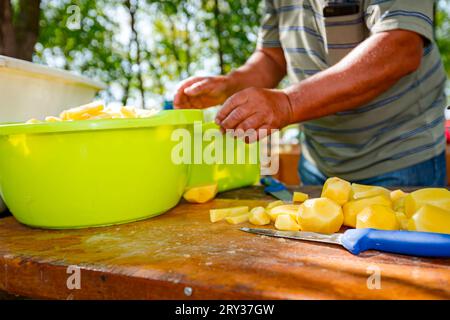 Ein älterer Bauernkocher schneidet Kartoffeln mit einem Messer in Scheiben auf dem Holzbrett und bereitet sie für das Kochen im Freien zu. Die Schüsseln sind voll mit Gemüse Stockfoto