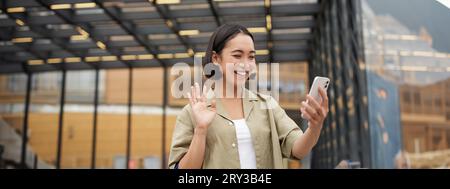 Glückliches asiatisches Mädchen winkt auf ihre Smartphone-Kamera, sagt Hallo zu einer Freundin im Video-Chat, ruft jemanden an, steht auf der Straße Stockfoto