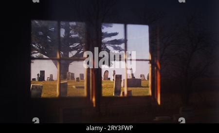 Alte Kiefer umgeben von Grabsteinen, eingerahmt durch ein antiquiertes Fenster einer alten Friedhofshütte. Stockfoto