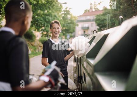 Glücklicher Teenager, der Plastikmüllbeutel in die Mülltonne bei Freunden steckt Stockfoto