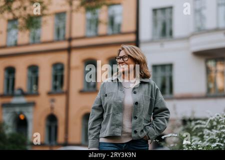 Glückliche Seniorin, die Jacke trägt, während sie vor dem Gebäude steht Stockfoto
