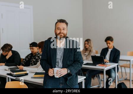 Porträt eines bärtigen, reiferen Professors, der eine Kaffeetasse hält und sich im Klassenzimmer auf den Schreibtisch lehnt Stockfoto